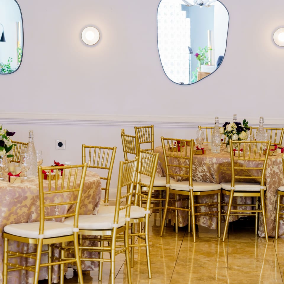 Elegant restaurant dining room with gold chairs, patterned tablecloths, floral centerpieces, glassware, and decorative wall mirrors.
