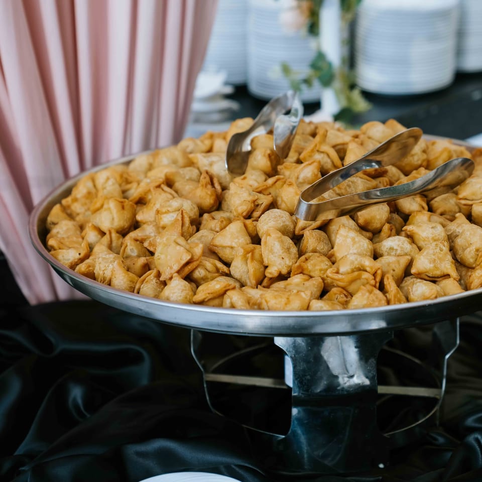 Large serving tray of golden fried pastries with metal tongs on a buffet table, beside a pink striped vase.