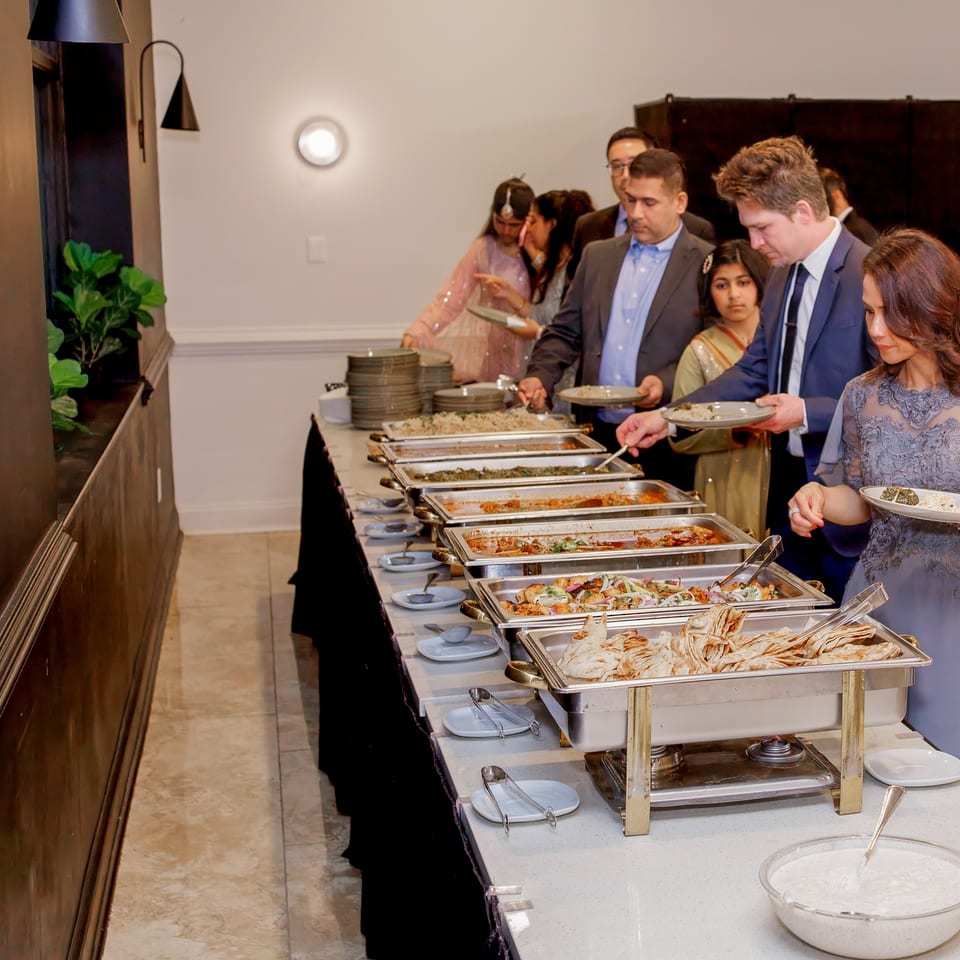 Guests serve themselves from a buffet of rice, vegetables, sauces, and flatbreads at an elegant indoor event.