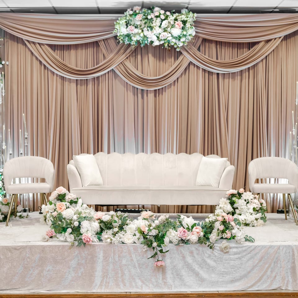 Elegant event stage with white loveseat, matching chairs, draped champagne curtains, candlelight, and abundant white and blush floral arrangements.