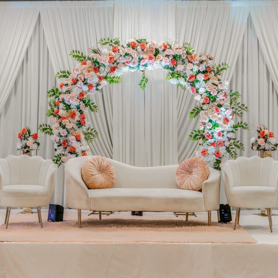 Elegant event stage with white drapery, floral arch, matching flower arrangements, and cream seating.