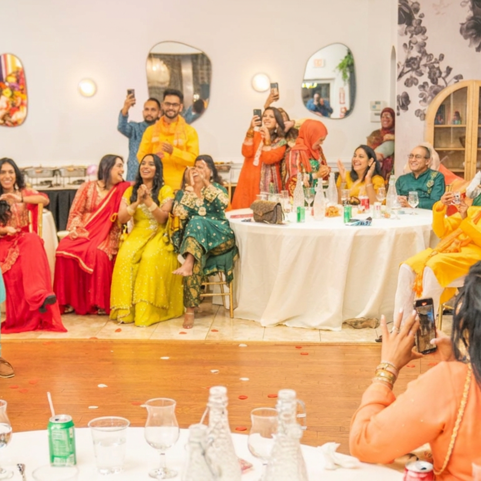 Guests in colorful Indian attire celebrate at a wedding reception while a bride in red and gold sits beside a decorated table.