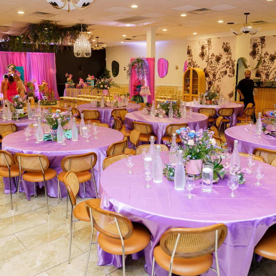 Vibrant restaurant event space with lavender-covered tables, floral centerpieces, gold chairs, and pink floral decorations.