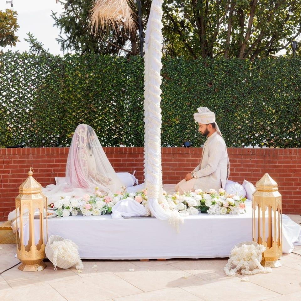Bride and groom sit beneath a floral wedding canopy on a decorated outdoor platform, framed by lanterns and sparklers.