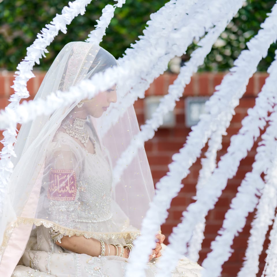 Bride in an embroidered veil and traditional attire sits behind cascading white floral decorations against a brick wall.