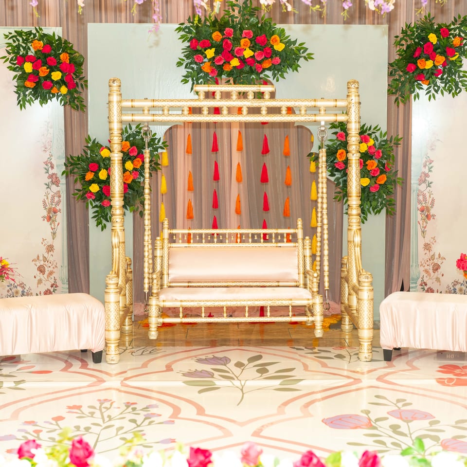 Ornate gold swing surrounded by colorful rose arrangements, lanterns, floral panels, and draped seating at a festive event.